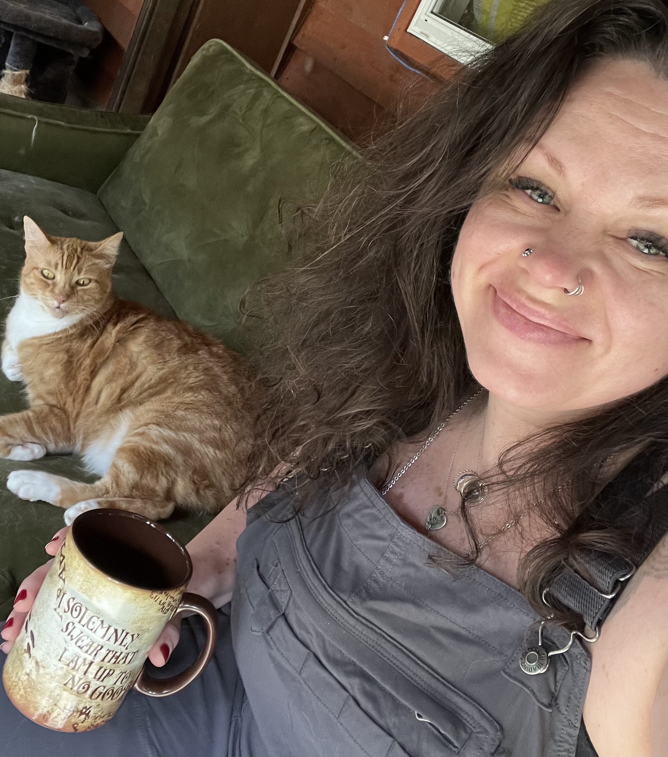 picture of Genevieve smiling and sitting on a green couch with an orange tabby cat holding a Harry Potter coffee mug that says I solemnly swear I am up to no good.